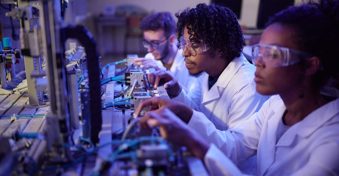 Three scientists in safety glasses and white coats work on electrical machinery